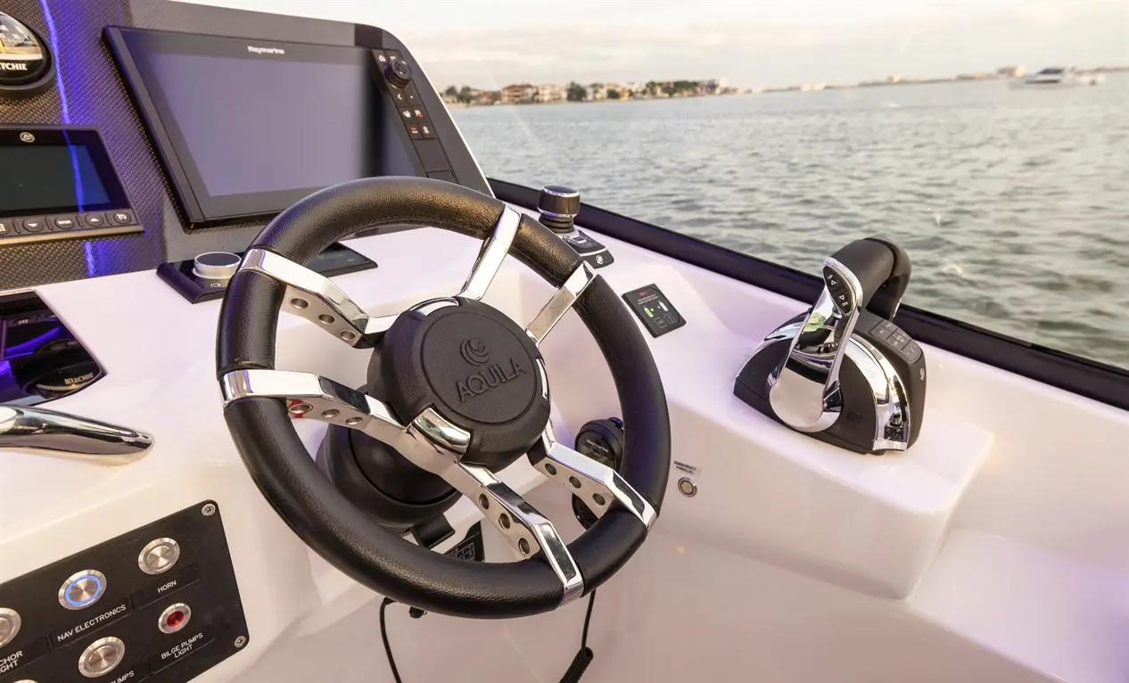Boat dashboard with an Aquila steering wheel, navigation screens, throttle controls, and water in the background.